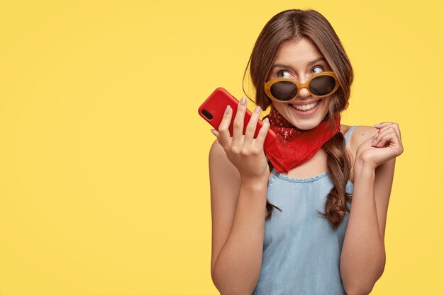 Pleased delightful female wears trendy shades, holds mobile phone, listens pleasant melody, enjoys high speed internet, waits for important call, isolated over yellow wall with copy space