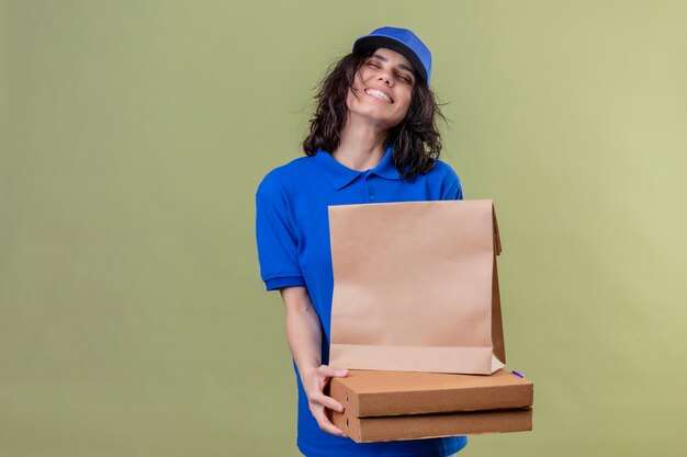 Ragazza piacevole di consegna in scatole della pizza della tenuta dell'uniforme blu e pacchetto di carta con gli occhi chiusi che sorridono con il fronte felice sopra la parete verde oliva di colore