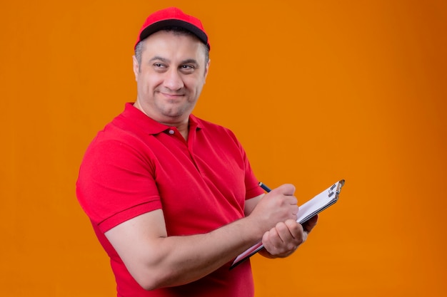 Pleased delivery man wearing red uniform and cap holding clipboard writing something looking aside with smile