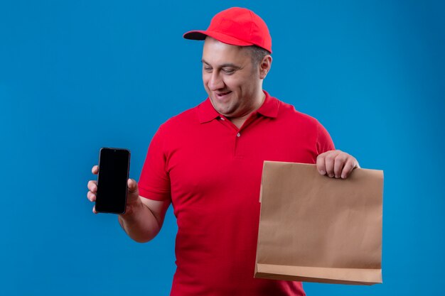 행복 한 얼굴로 웃 고 휴대 전화를 보여주는 빨간 유니폼과 모자 들고 종이 패키지를 입고 기쁘게 배달 남자
