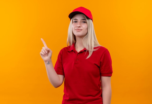 孤立したオレンジ色の背景に赤いTシャツとキャップポイントを身に着けている幸せな配達の若い女の子