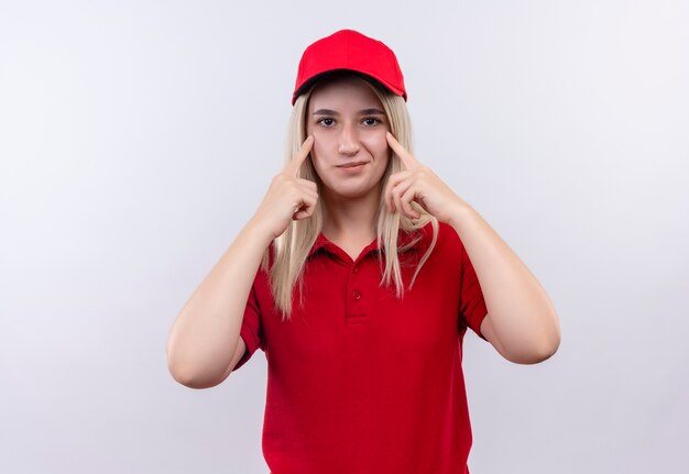 Довольная молодая девушка доставки в красной футболке и кепке положила пальцы на глаза на изолированном белом фоне