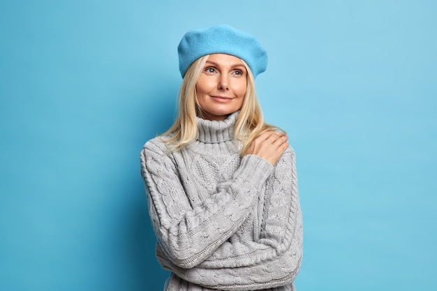 pleased dreamy blonde European woman has gentle satisfied expression wears beret and knitted jumper concentrated into distance with pensive look.