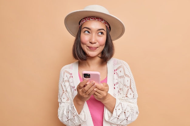 Довольная мечтательная молодая азиатская женщина держит в руках современный смартфон, использует мобильное приложение для онлайн-чата, смотрит в сторону в стильной шляпе, белая вязаная шаль, изолированная на коричневой стене