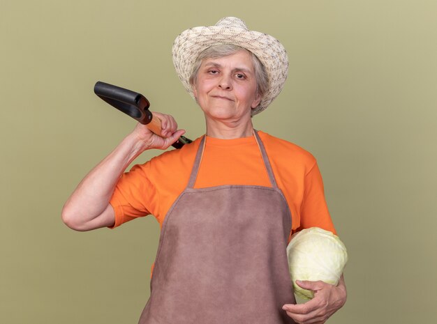 Pleased elderly female gardener wearing gardening hat holding cabbage and spade on shoulder