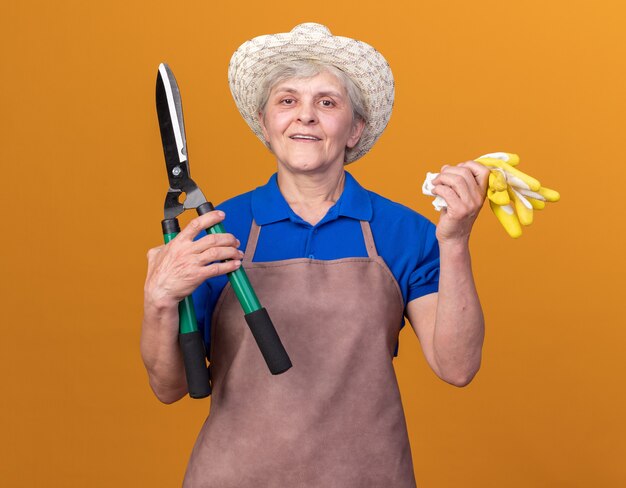 Pleased elderly female gardener wearing gardening hat holding gardening scissors and gloves on orange