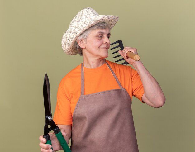 원예 가위와 갈퀴를 들고 원예 모자를 쓰고 기쁘게 노인 여성 정원사 올리브 그린에 측면을보고