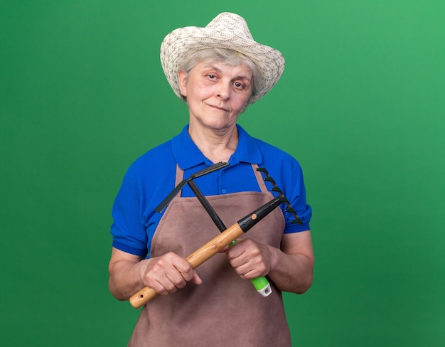 Pleased elderly female gardener wearing gardening hat holding rake and hoe rake on green