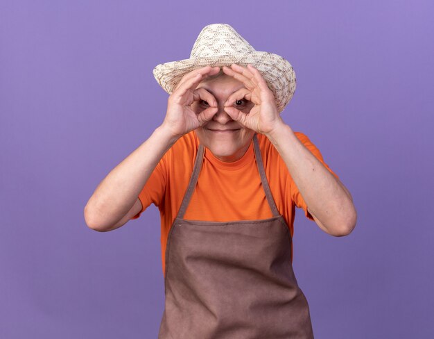 Pleased elderly female gardener wearing gardening hat through fingers isolated on purple wall with copy space