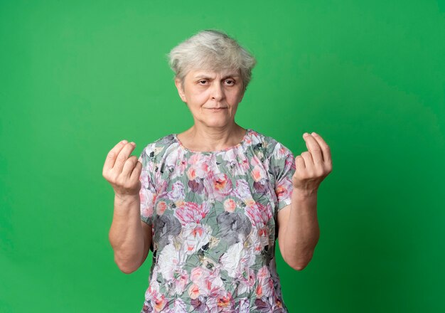 Pleased elderly woman gestures money hand sign looking isolated on green wall