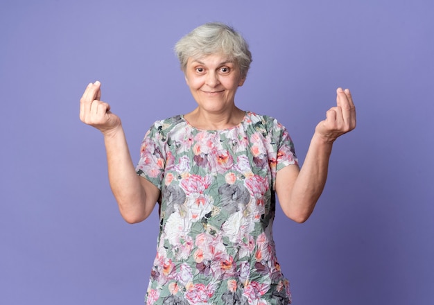 Free photo pleased elderly woman gestures money hand sign with two hands isolated on purple wall