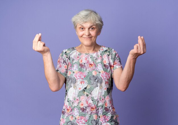 Довольная пожилая женщина жестикулирует знак рукой деньги двумя руками, изолированными на фиолетовой стене