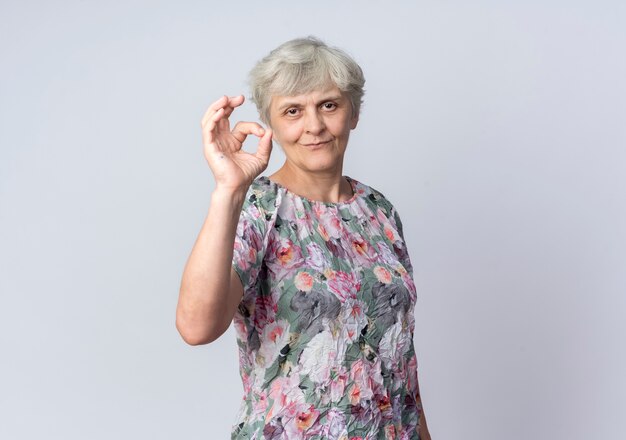 Pleased elderly woman gestures ok hand sign isolated on white wall