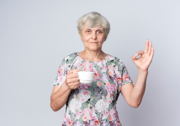 Pleased elderly woman holds cup and gestures ok hand sign isolated on white wall