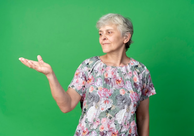 Pleased elderly woman looks at empty hand isolated on green wall