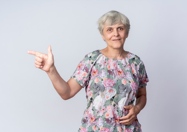 Pleased elderly woman points at side isolated on white wall