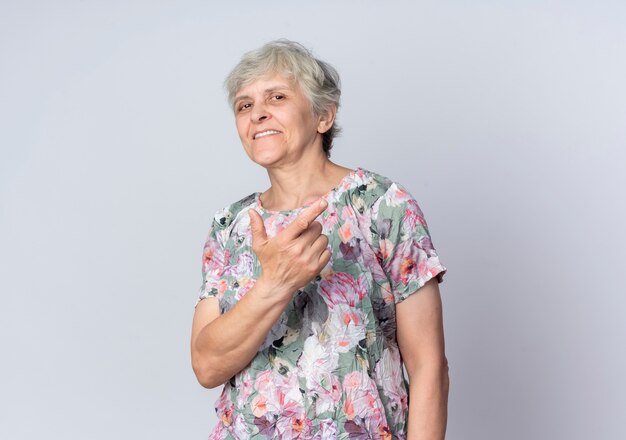 Pleased elderly woman points at side looking isolated on white wall