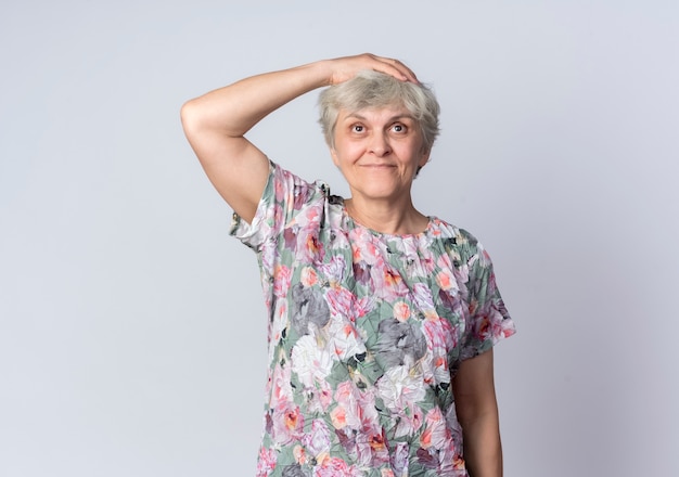 Pleased elderly woman puts hand on head isolated on white wall