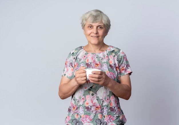 La donna anziana lieta sta tenendo la tazza isolata sulla parete bianca