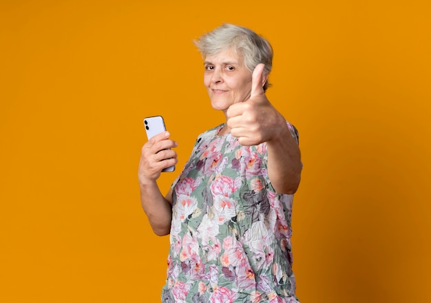 Free photo pleased elderly woman stands sideways holding phone and thumbs up isolated on orange wall