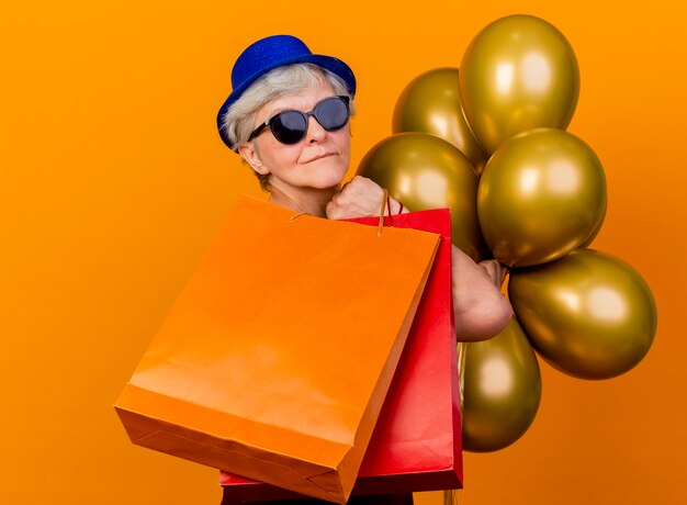 パーティーハットを身に着けているサングラスの満足している年配の女性は、コピースペースでオレンジ色の壁に隔離されたヘリウム風船と紙の買い物袋を持って横に立っています