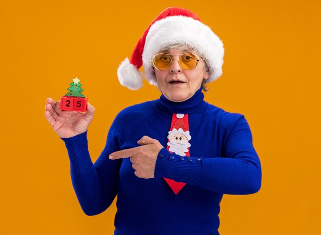 Pleased elderly woman in sun glasses with santa hat and santa tie holding and pointing at christmas tree ornament isolated on orange wall with copy space