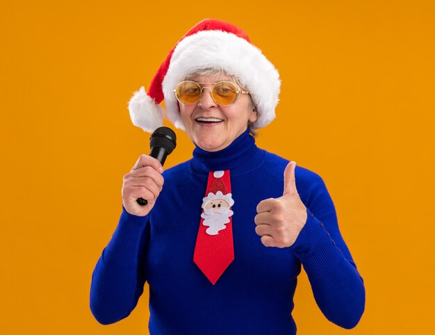 Pleased elderly woman in sun glasses with santa hat and santa tie holds mic and thumbs up isolated on orange wall with copy space