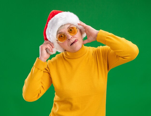 Довольная пожилая женщина в солнцезащитных очках в шляпе санта-клауса высунула язык на зеленой стене с копией пространства