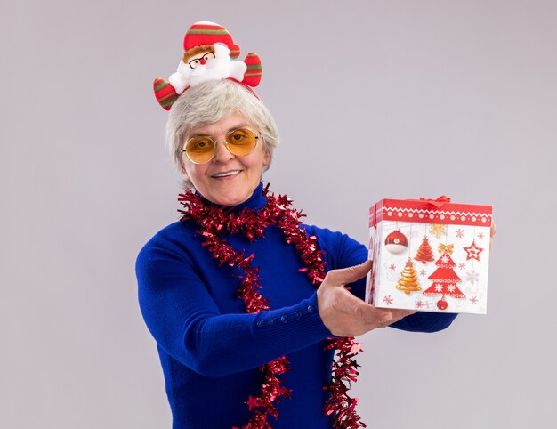 Pleased elderly woman in sun glasses with santa headband and garland around neck holds christmas gift box isolated on white wall with copy space