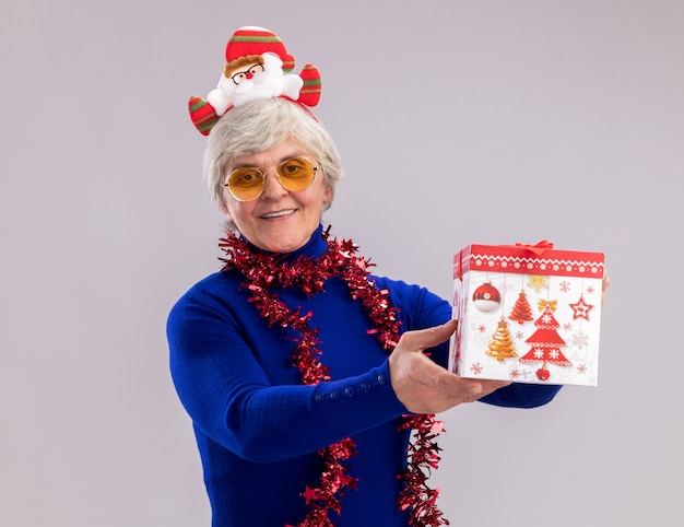 Pleased elderly woman in sun glasses with santa headband and garland around neck holds christmas gift box isolated on white wall with copy space