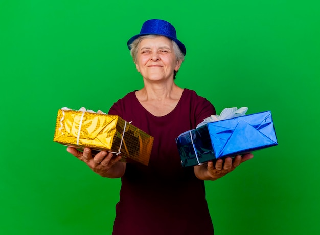 Pleased elderly woman wearing party hat holds gift boxes on green