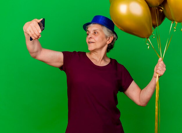 Pleased elderly woman wearing party hat holds helium balloons looking at phone on green