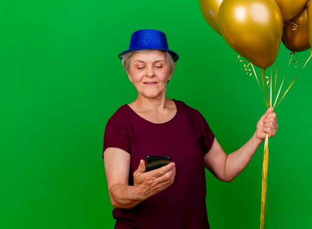 Pleased elderly woman wearing party hat holds helium balloons and looks at phone on green