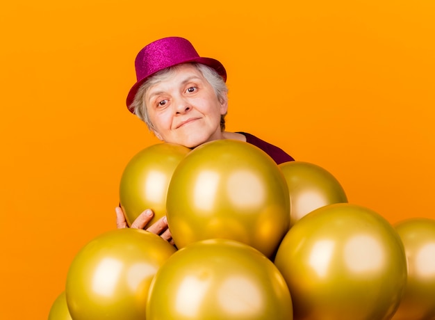 Felice donna anziana che indossa cappello da festa tiene e sta con palloncini di elio su arancione