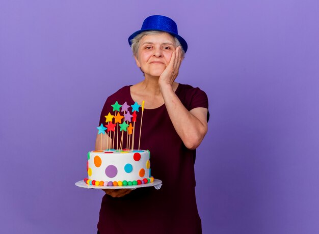 Довольная пожилая женщина в шляпе для вечеринки кладет руку на лицо и держит торт ко дню рождения, изолированный на фиолетовой стене