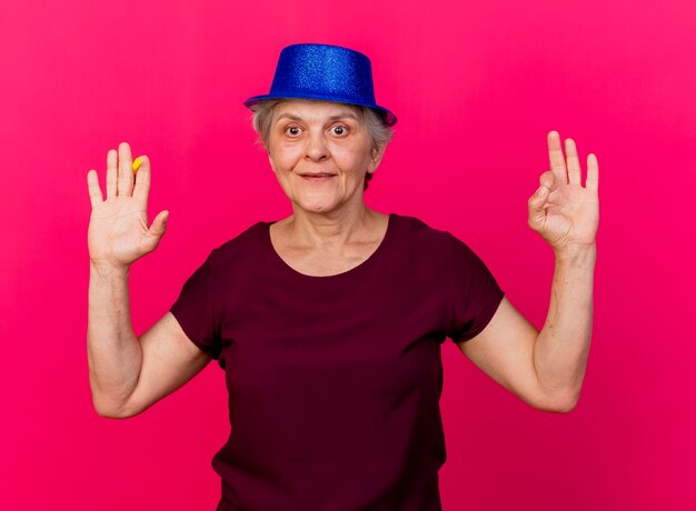 Pleased elderly woman wearing party hat stands with raised hand holding whistle and gesturing ok hand sign isolated on pink wall