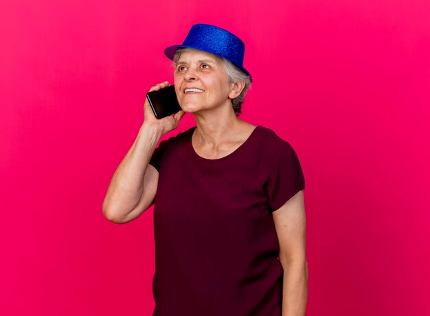 Pleased elderly woman wearing party hat talks on phone on pink