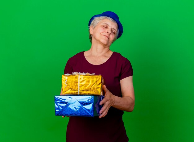 Pleased elderly woman wearing party hat with closed eyes holds gift boxes on green