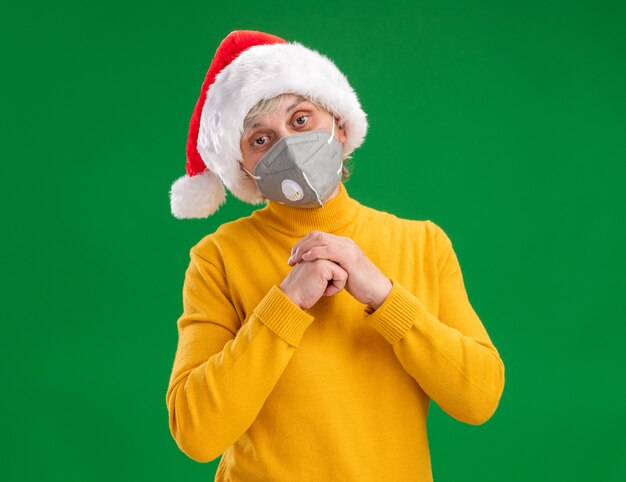 Pleased elderly woman with santa hat wearing medical mask holding hands together looking at camera isolated on green background with copy space