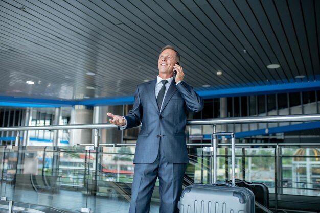 Довольный элегантный бизнесмен средних лет с багажом разговаривает по смартфону в терминале аэропорта