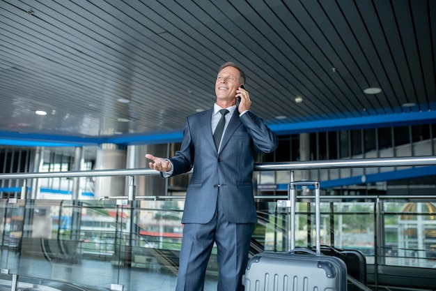 Pleased elegant middle-aged businessman with the luggage talking on the smartphone at the airport terminal