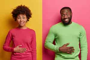 Free photo pleased ethnic young woman and man keep hands on stomach, feel satiety after eating tasty nutritious dinner, smile positively, happy not to be hungry, pose against yellow and pink wall