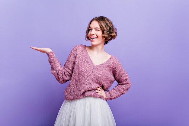 Pleased european woman in trendy white skirt funny posing  Attractive caucasian female model with short curly hair standing on purple wall in stylish sweater.