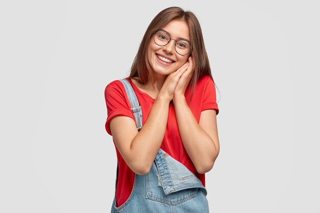 Pleased European young woman with cheerful expression, keeps both hands near face, being touched by compliment