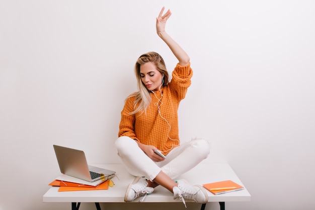 Pleased fair-haired woman in trendy sport shoes listening music in earphones and looking at laptop screen