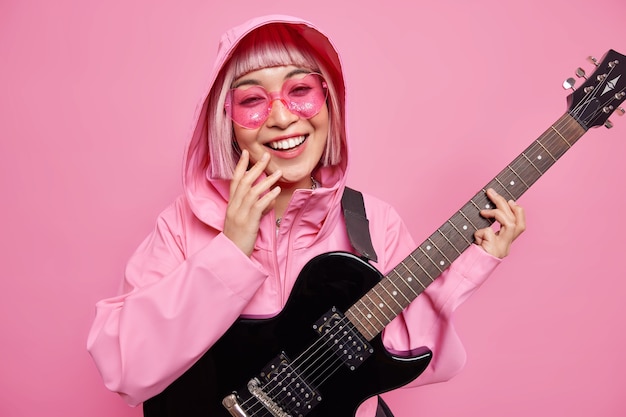 Foto gratuita la donna alla moda soddisfatta vestita con gli occhiali da sole rosa della giacca alla moda si diverte finge di essere una rock star sorride ampiamente usa la chitarra elettrica