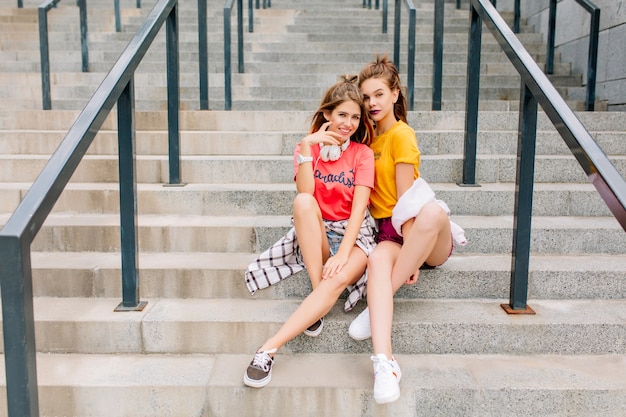 Pleased female friends chilling together on stone staircase with legs crossed posing emotionally