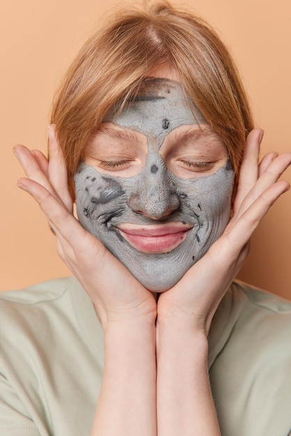 Pleased female model applies nourishing clay mask holds palms over face smiles gently keeps eyes closed undergoes beauty procedures at home isolated over beige background Facial treatments