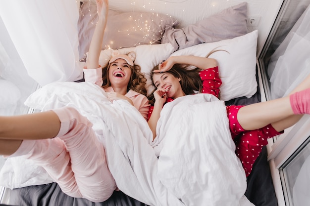 Pleased female model lying under white blanket and laughing. Indoor shot of two cheerful girls spending weekend morning in bed.