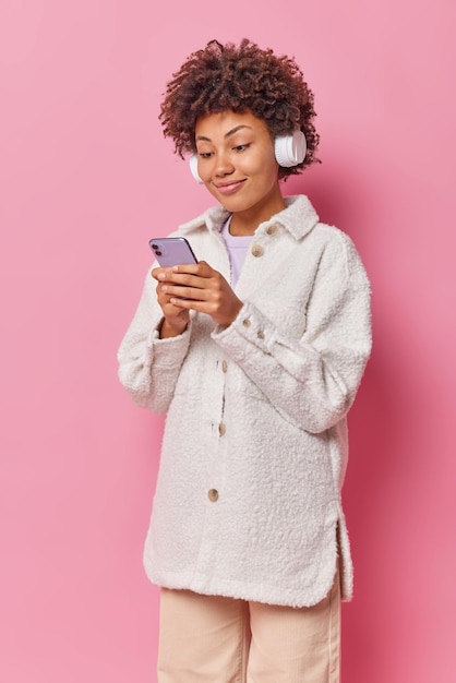 Pleased female model with curly hair concentrated at smartphone screen downloads songs to playlist wears stereo headphones on ears isolated over pink wall